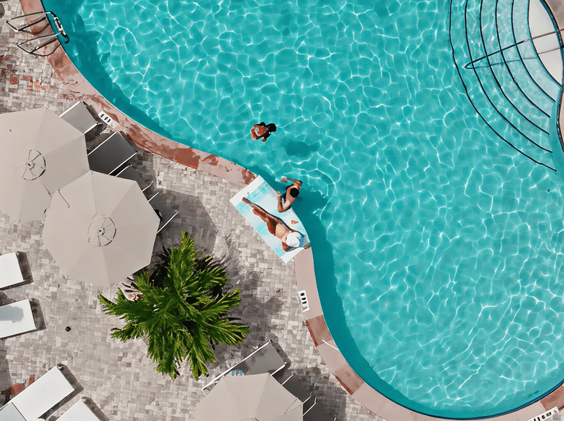 Aerial view of two people near the edge of a clear blue swimming pool; one is sunbathing on a lounger, the other stands nearby. White umbrellas, loungers, and a palm tree surround the pool on stone tiles.