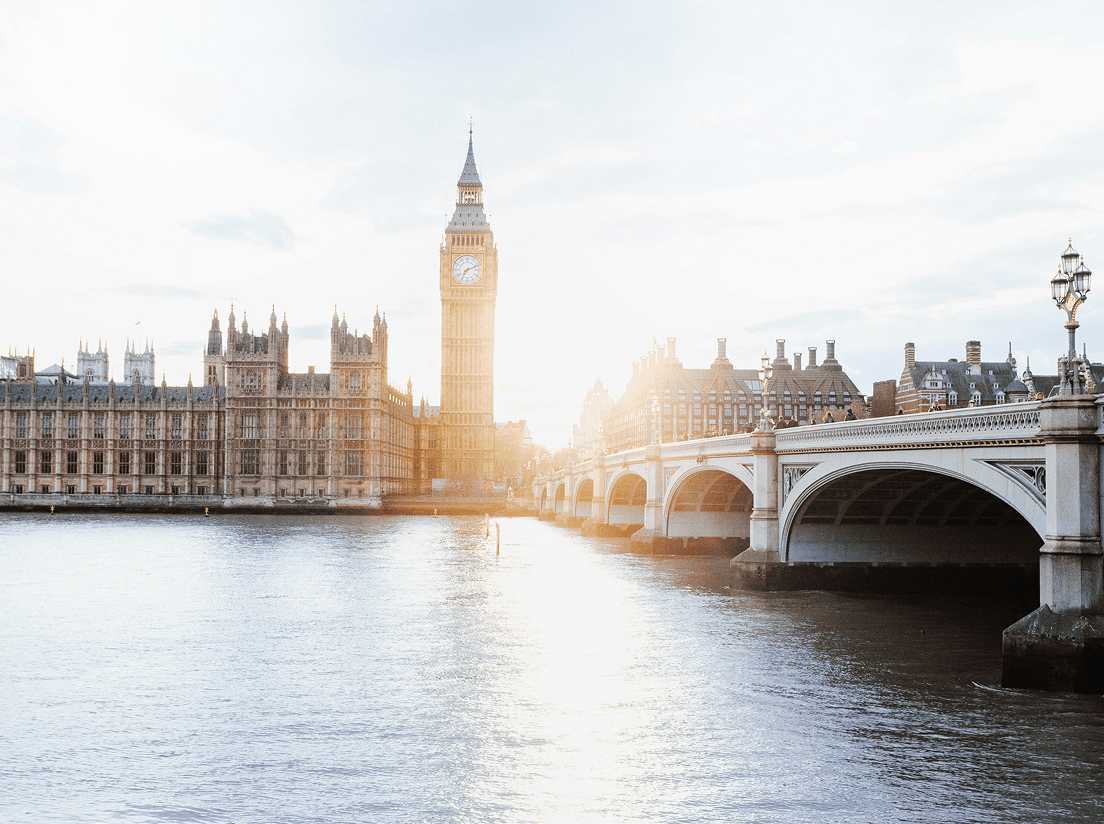 The sun shines brightly behind Big Ben and the Houses of Parliament in London, with Westminster Bridge crossing the River Thames in the foreground.