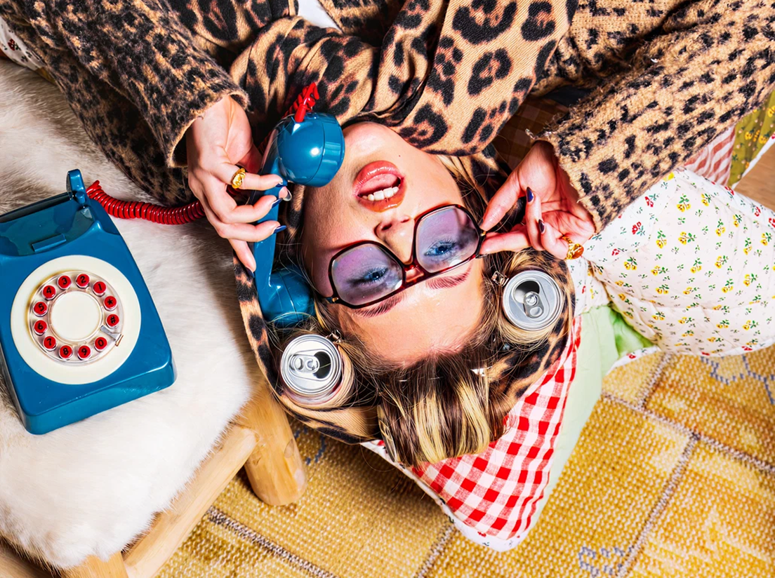 A woman wearing leopard print and large glasses lies upside down on a couch, talking on a blue rotary phone. She has soda cans in her hair like curlers and colorful pillows beneath her head.