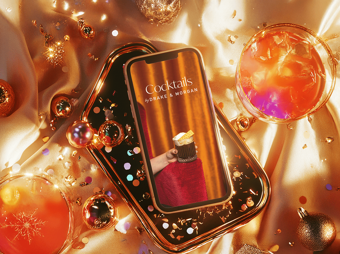 A smartphone displaying a "Cocktails by Drake & Morgan" screen rests on a festive tray surrounded by gold ornaments, confetti, and two glasses of orange cocktail, all atop shimmering golden fabric.