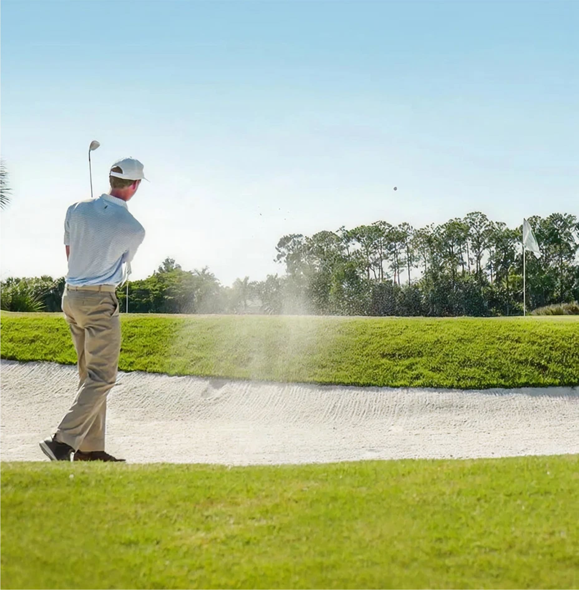 A golfer in a white shirt and khaki pants swings a club from a sand bunker, sending sand into the air on a golf course with a green flag in the background and trees lining the horizon.