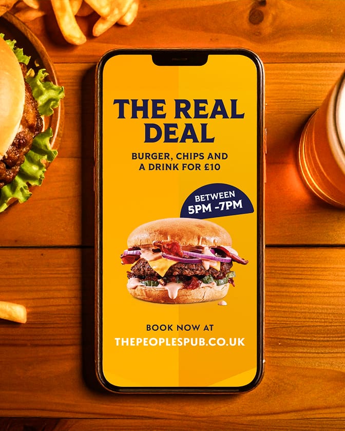 A smartphone on a wooden table displays an ad: “THE REAL DEAL—Burger, chips and a drink for £10, between 5pm–7pm.” Next to the phone are fries, a burger, and a glass of beer.