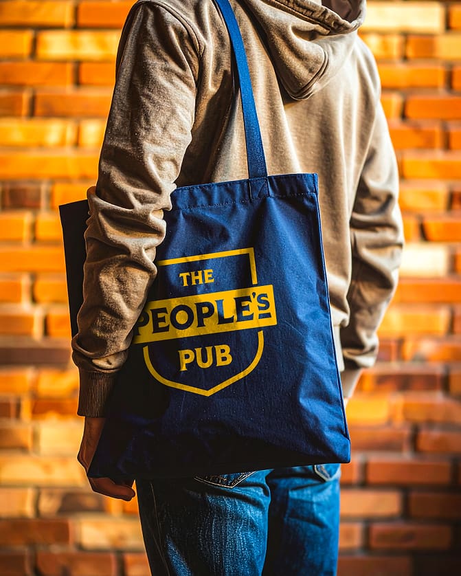 A person in a brown hoodie and jeans stands in front of a brick wall, holding a blue tote bag with yellow text that reads "THE PEOPLE'S PUB.