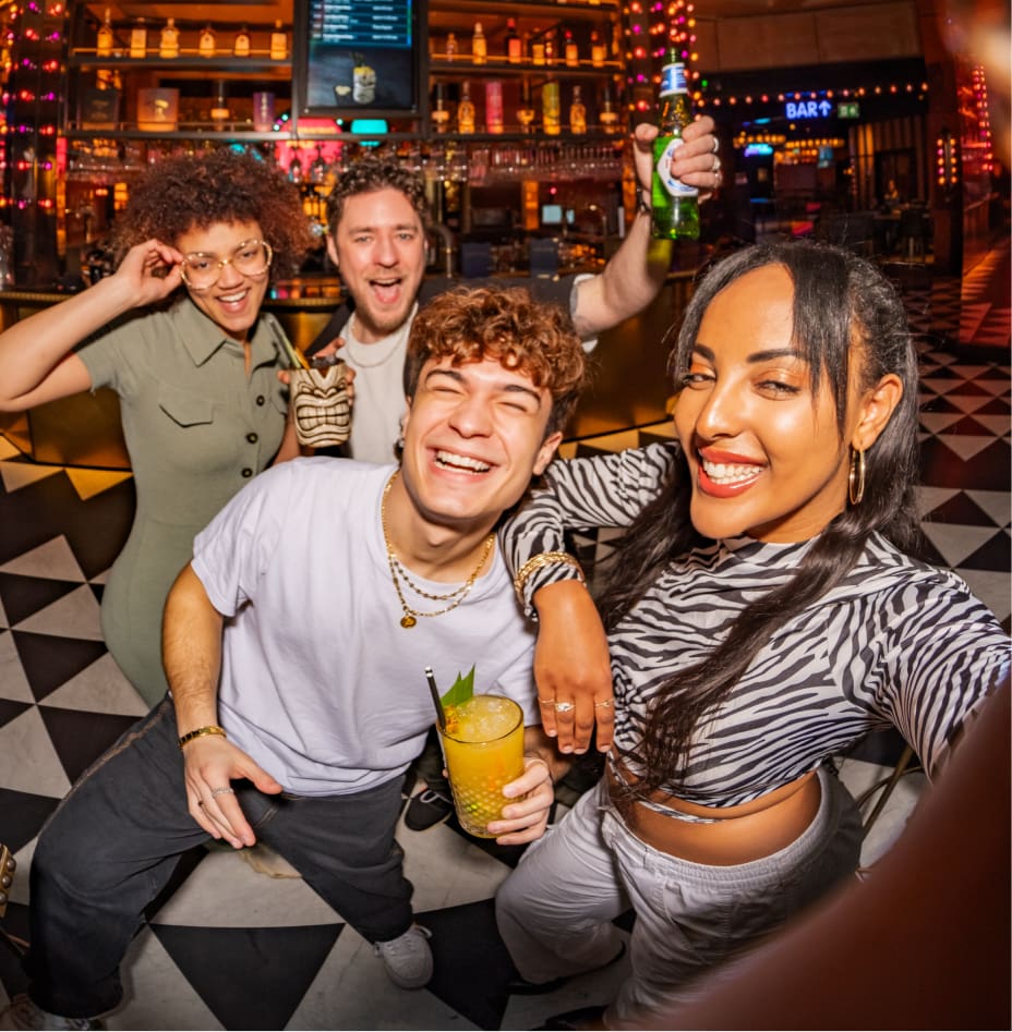 Four friends are smiling and posing for a selfie at a lively bar with colorful lights and patterned flooring. They are holding drinks, looking happy and energetic.