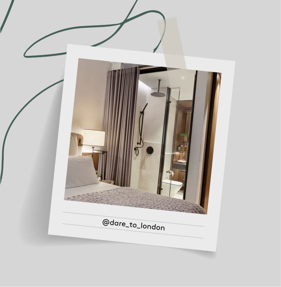 A modern hotel room featuring a neatly made bed, a bedside lamp, and a glass-enclosed shower with black fixtures. The scene is framed like a Polaroid with "@dare_to_london" written below.