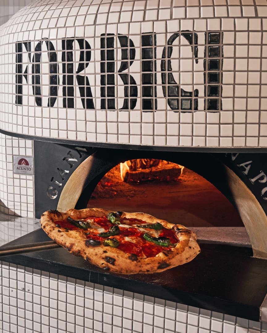 A wood-fired pizza oven labeled "FORBICI" with a pizza topped with tomatoes, basil, and melted cheese being removed from the oven on a peel, flames visible inside.