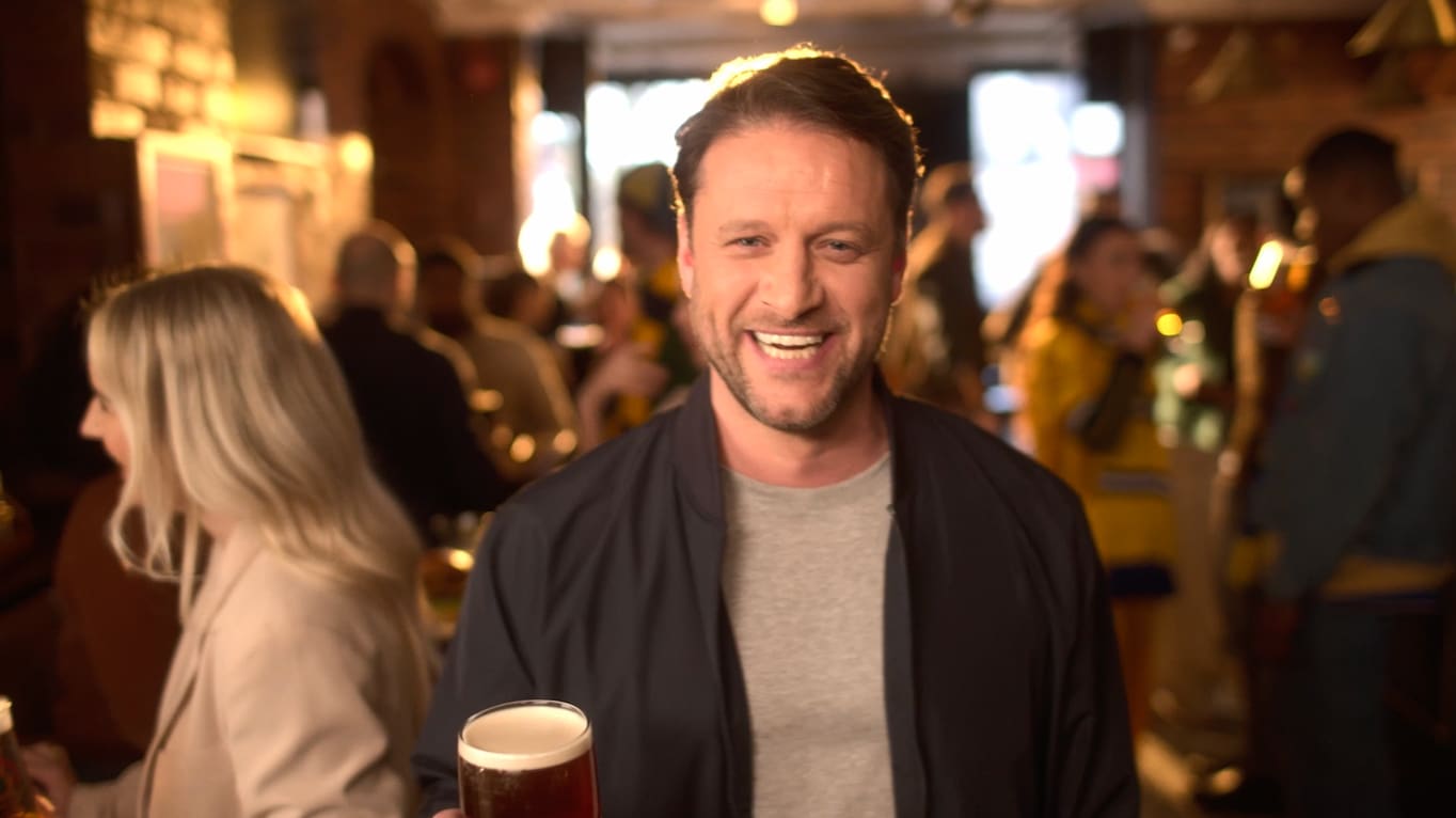A smiling man in a dimly lit bar holds a pint of beer, with people socializing and mingling in the blurred background.