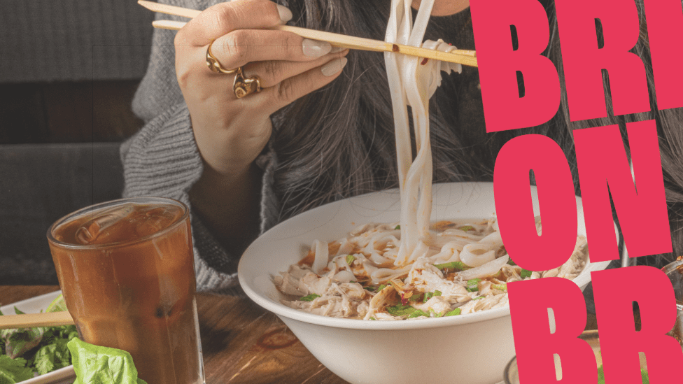 A person eating noodle soup with chopsticks next to a glass of iced tea on a wooden table, with bold pink letters partially covering the right side of the image.