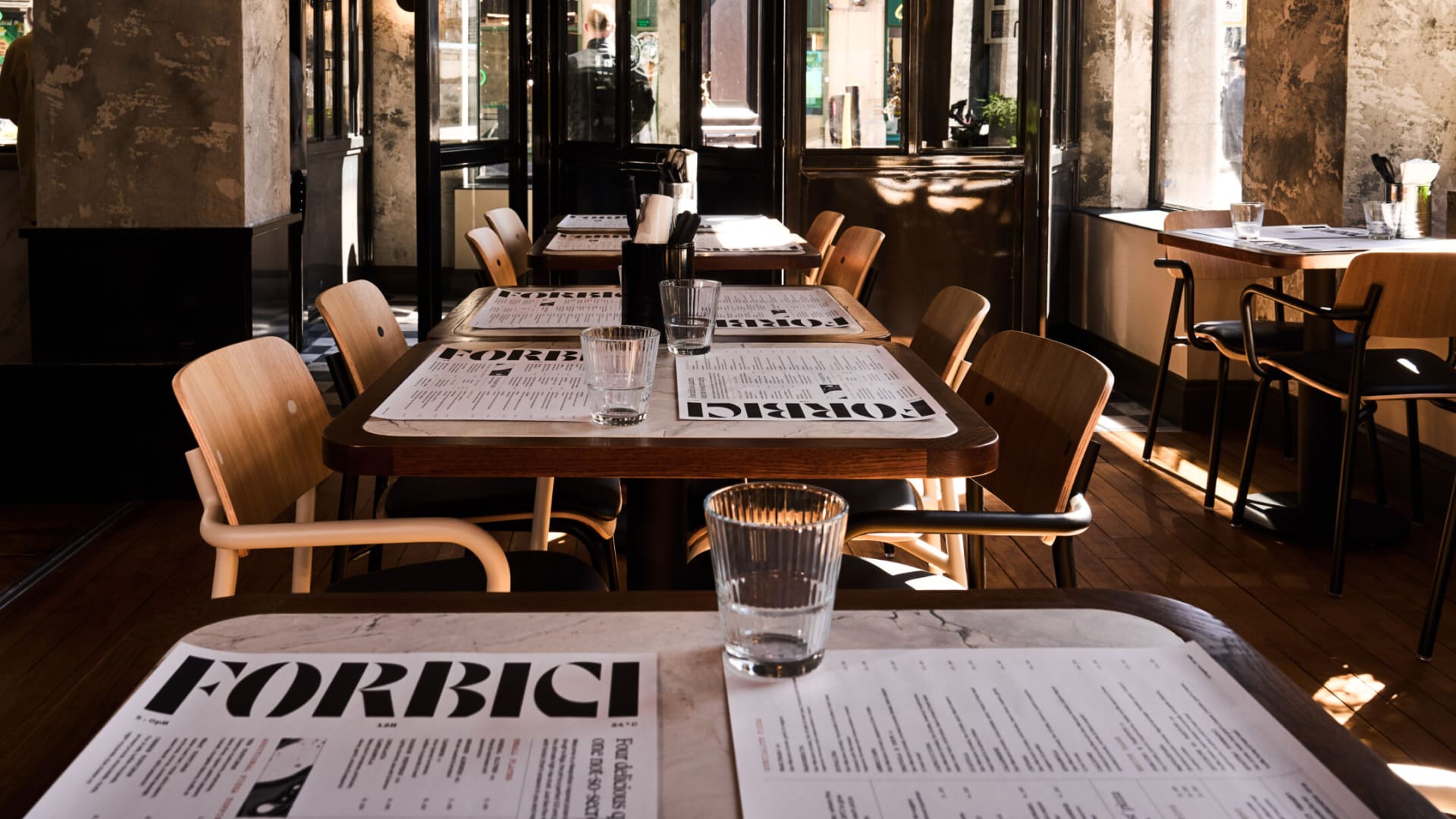 Interior of a modern restaurant with wooden tables set with menus labeled “FORBICI,” drinking glasses, and chairs. Sunlight streams in through large windows, creating a warm and inviting atmosphere.
