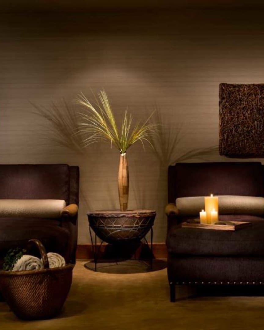 Two dark armchairs face each other with a woven basket of blankets on the left. In the center, a vase with tall grass decor sits on a table, and two lit candles rest on the right armchair's armrest, creating a cozy ambiance.