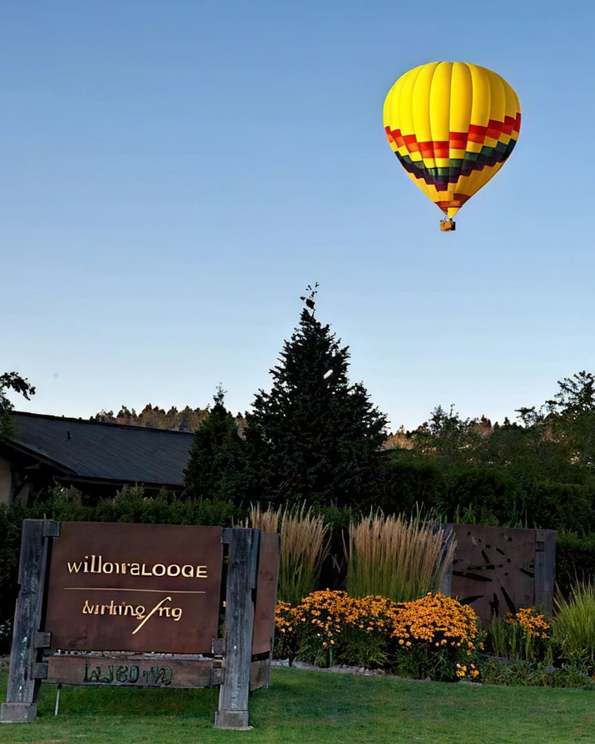 A colorful hot air balloon floats in a clear sky above a lodge surrounded by greenery and yellow flowers, with a wooden sign in the foreground reading "Willow Lodge Parking.
