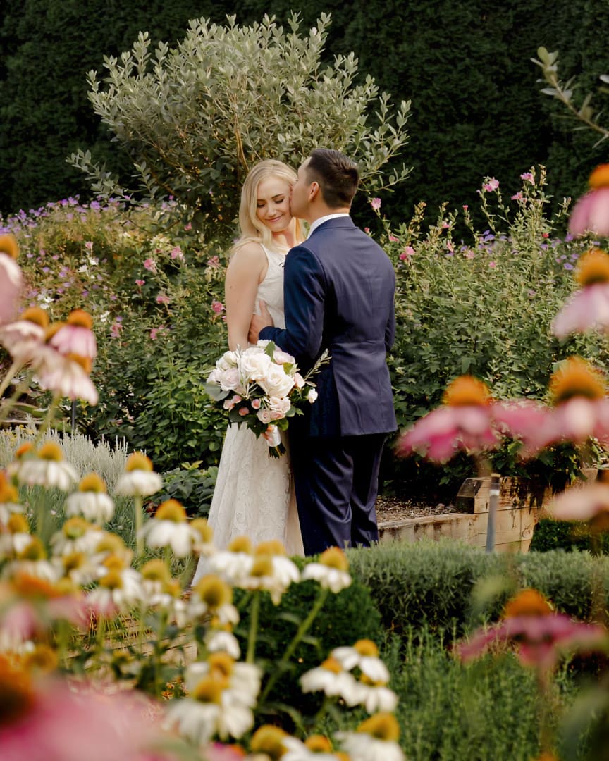 A bride and groom stand close together in a lush garden, surrounded by blooming flowers. The groom kisses the bride’s forehead as she smiles, holding a bouquet of white and pink flowers.