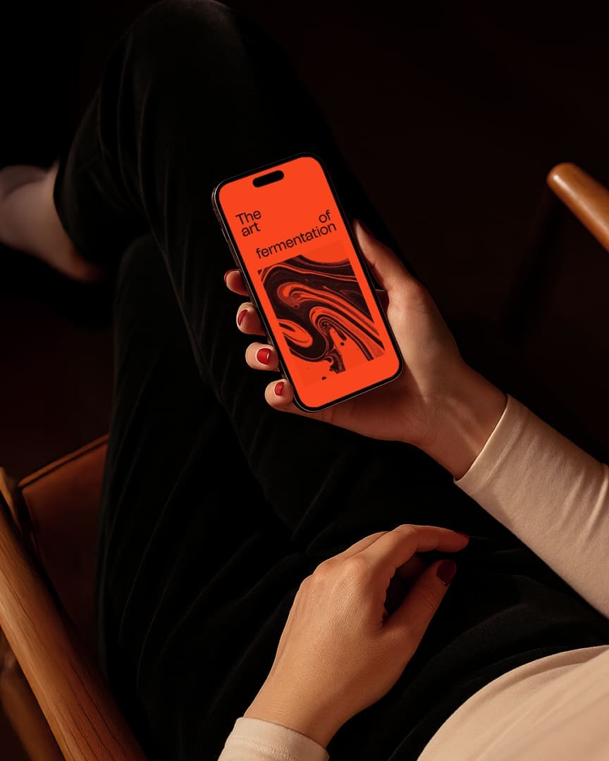 A person with red-painted nails holds a smartphone displaying an orange screen with abstract black swirls and the text “The art of fermentation” while sitting in a chair with legs crossed.