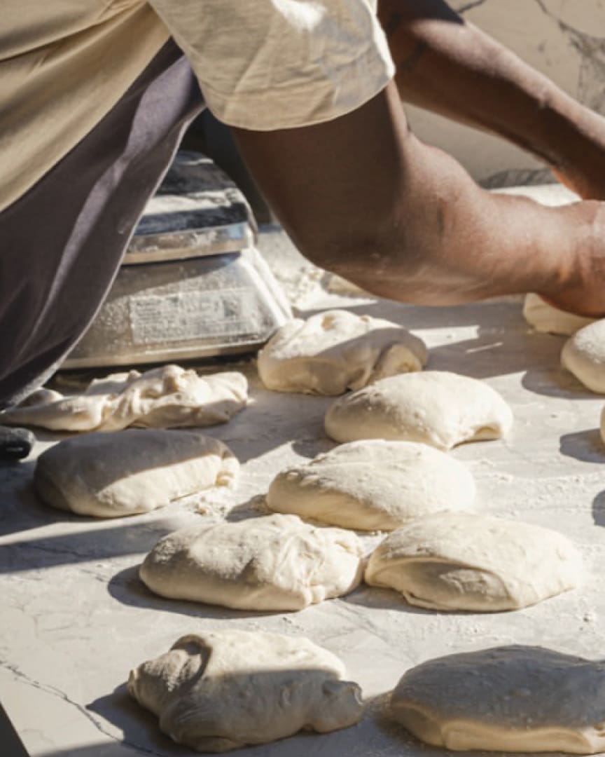 A person works with multiple pieces of raw bread dough on a floured surface, preparing them for baking. Their arms are visible as they handle the dough. A scale is in the background.