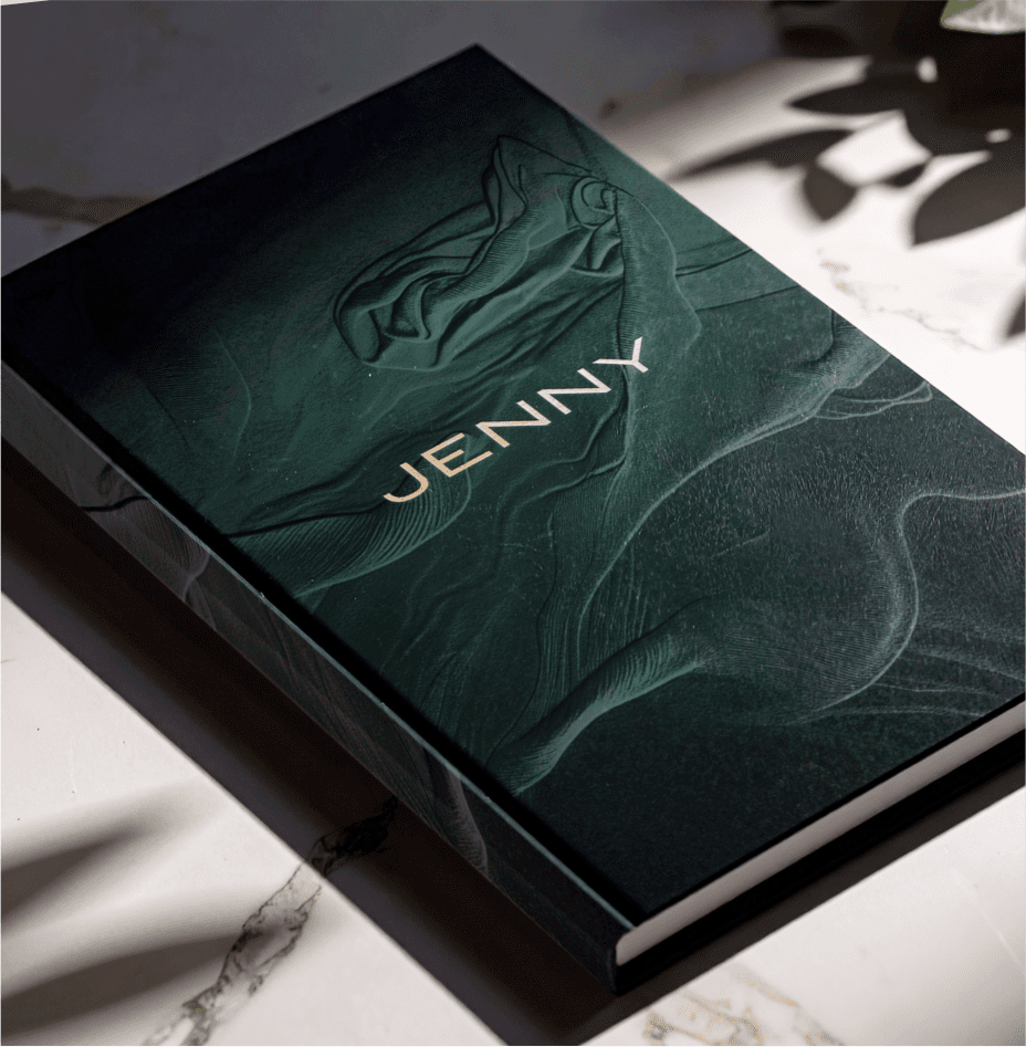 A hardcover book with a textured dark green cover and the word "JENNY" in gold letters lies on a white marble surface, partially shaded by plant leaves.