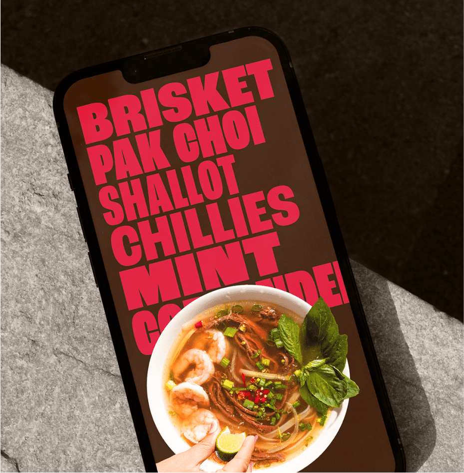 A smartphone displaying large red text listing brisket, pak choi, shallot, chillies, and mint, with a bowl of noodle soup topped with herbs, shrimp, and lime at the bottom of the screen.