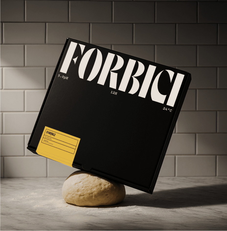A black pizza box labeled "FORBICI" balances on a ball of dough on a marble surface, with white subway tiles as the background. A yellow sticker is on the lower left corner of the box.