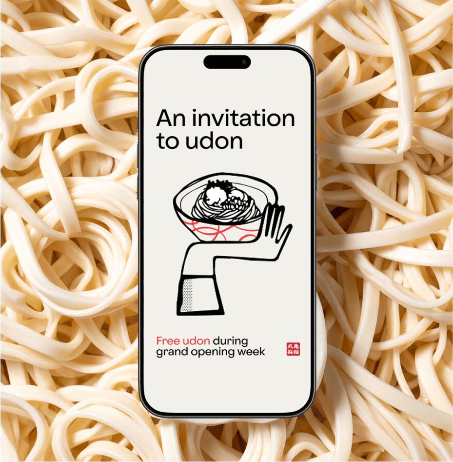A smartphone displays an invitation for free udon during grand opening week, featuring an illustration of a hand holding a bowl of udon. The phone is placed on a background of uncooked udon noodles.