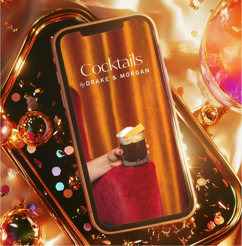A smartphone displays a cocktail menu titled "Cocktails by Drake & Morgan," with a hand holding a fancy drink. The phone rests on a festive, gold and confetti-covered surface surrounded by colorful drinks.