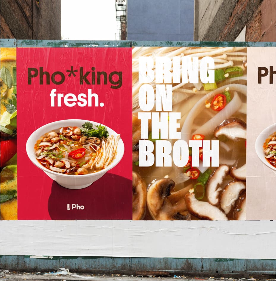 Posters on a brick wall advertise pho. The left poster says "Pho*king fresh." with a bowl of pho. The right poster says "BRING ON THE BROTH" over an image of pho with mushrooms, noodles, and red chili.