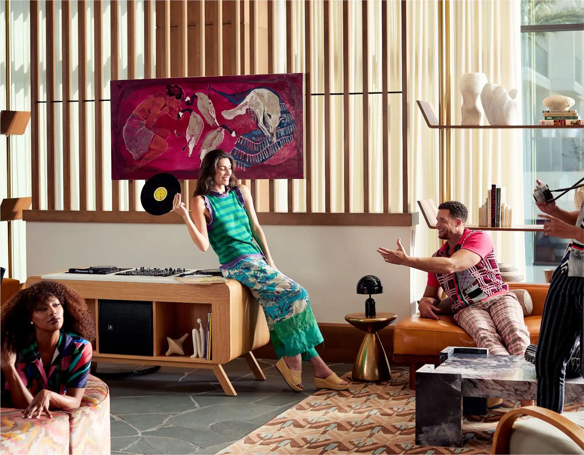 Four people relax in a stylish, modern living room. One woman sits on a wooden console by a turntable, holding a vinyl record. Others lounge and interact, surrounded by art, warm lighting, and mid-century furniture.