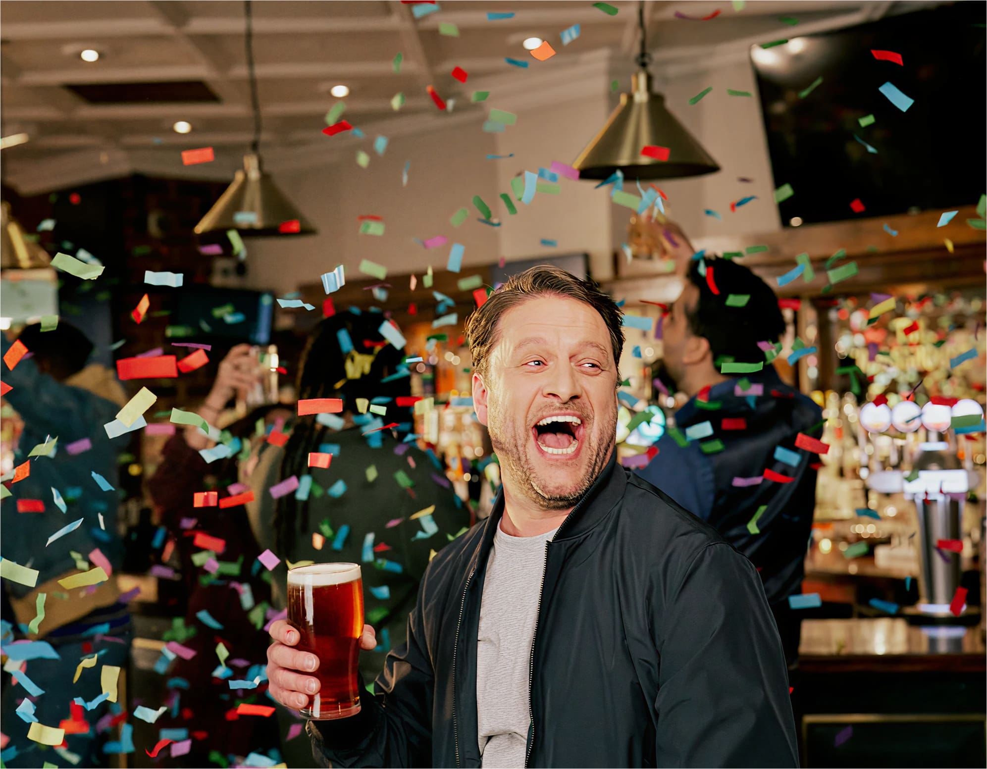 A man holding a pint of beer laughs joyfully as colorful confetti falls around him in a lively bar with people and lights in the background.