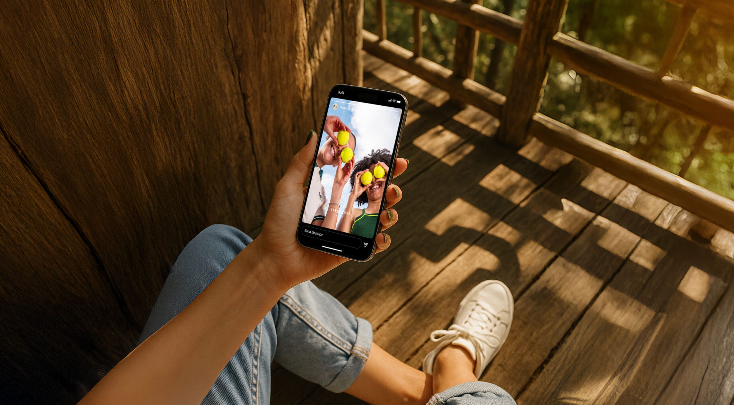 A person sitting on a wooden deck holds a smartphone showing a photo of three friends posing together, wearing sunglasses and smiling. The person’s hand and white sneaker are visible in the foreground.