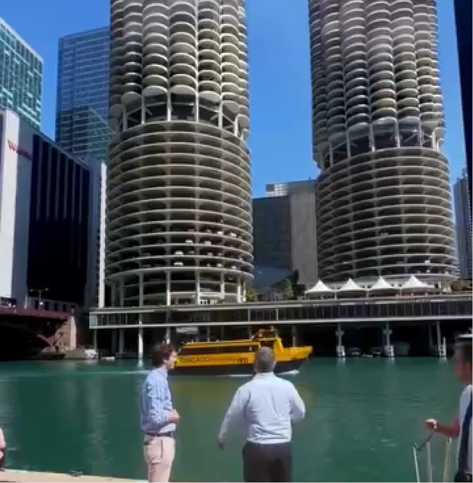 Three people stand by a river as a yellow tour boat passes by. Behind them are two round, uniquely designed high-rise buildings and other skyscrapers under a clear blue sky.