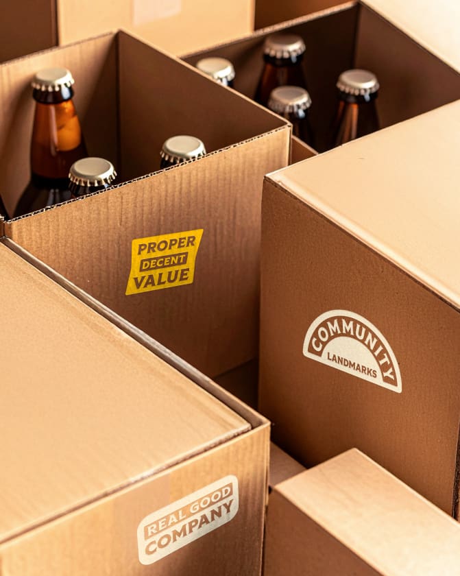 Several brown cardboard boxes filled with glass bottles with metal caps, some boxes labeled with phrases like "PROPER DECENT VALUE," "COMMUNITY LANDMARKS," and "REAL GOOD COMPANY.