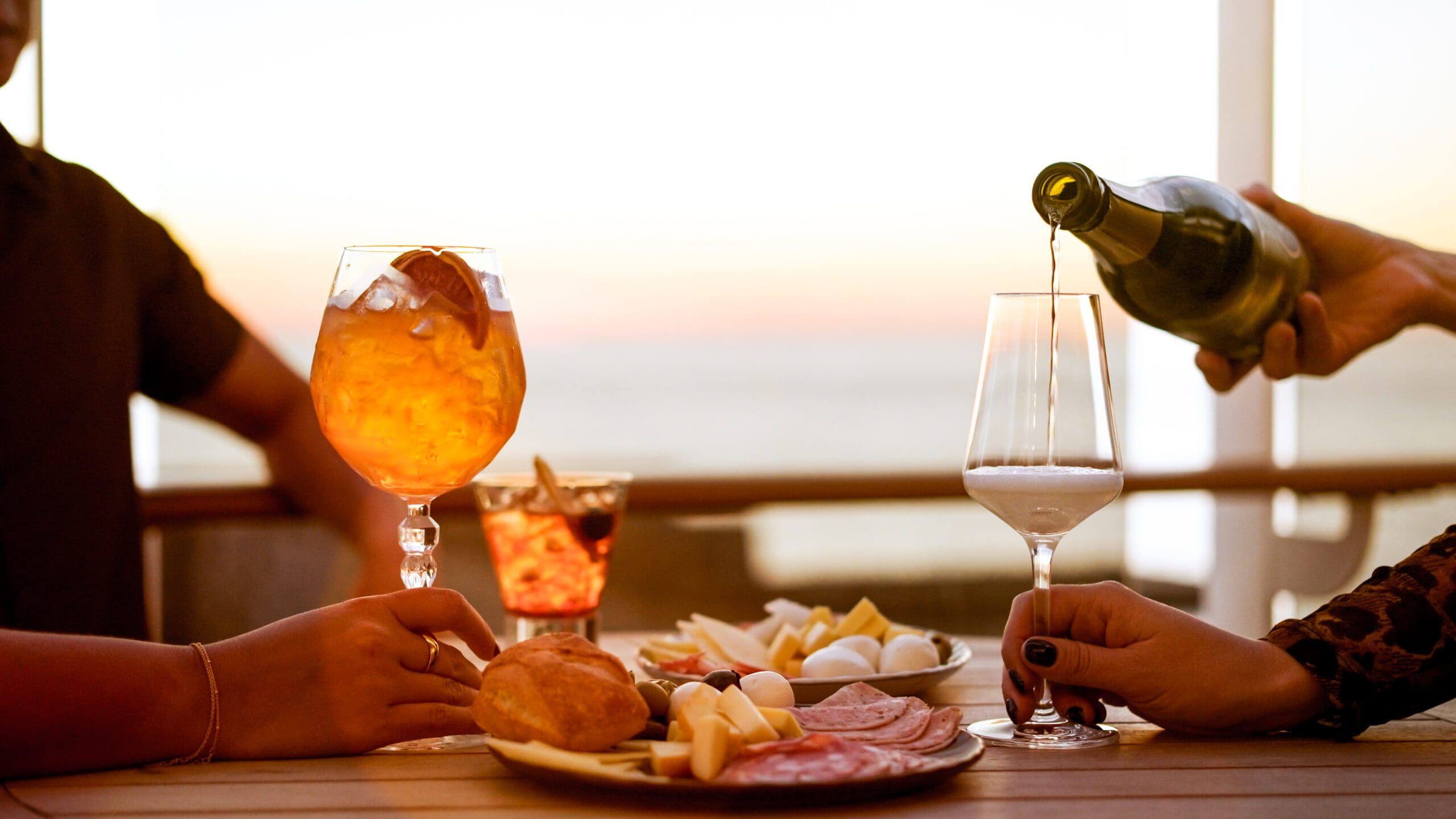 A close-up of a table with assorted cheeses, meats, bread, and drinks. One hand holds an orange cocktail, another pours white wine into a glass. The background shows a sunset over water.