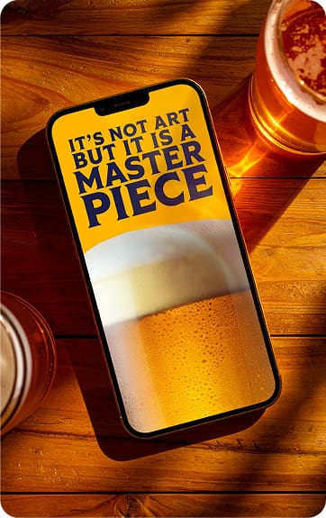 A smartphone on a wooden table displays an image of beer with foam and the text: "It's not art but it is a masterpiece." Two glasses of beer sit nearby, casting shadows.