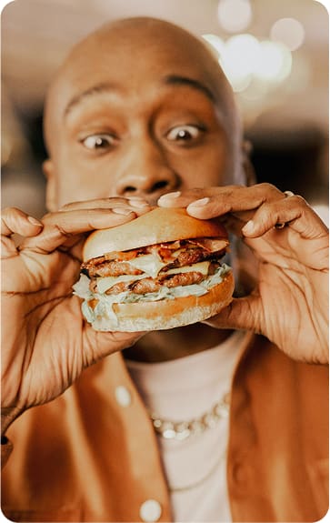 A person holding a large hamburger with lettuce, sauce, and bacon, looking excited and about to take a bite. The background is softly blurred.