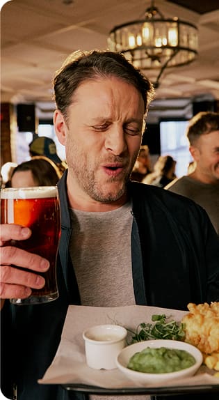 A man holding a pint of beer and a plate of food makes a funny face, as if reacting to the taste. He stands in a lively, warmly lit restaurant or pub with other people in the background.