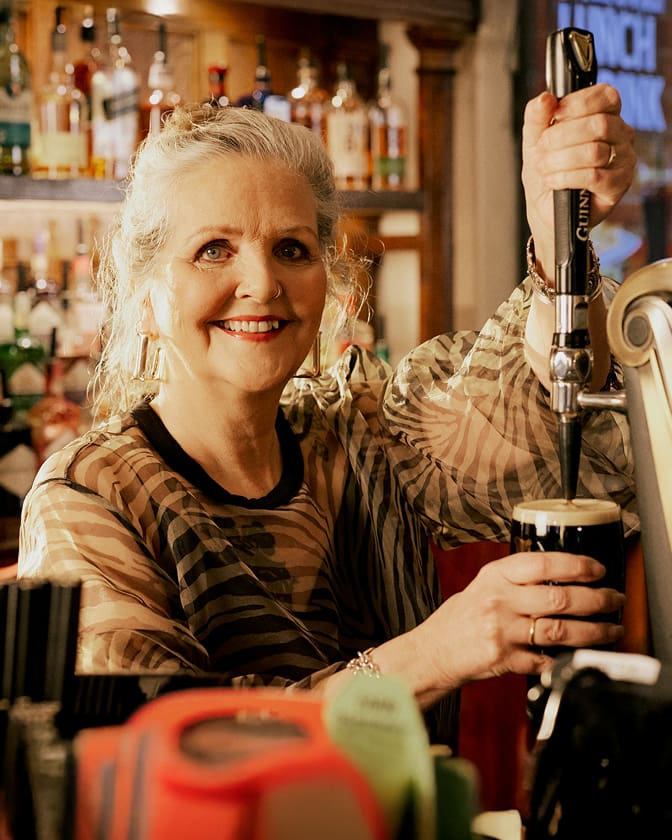 A smiling older woman with gray hair pours a pint of dark beer from a tap behind a bar, surrounded by bottles and bar equipment. She wears a sheer, striped top and large earrings.