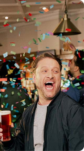 A man holding a pint of beer smiles widely while colorful confetti falls around him in a lively indoor setting.