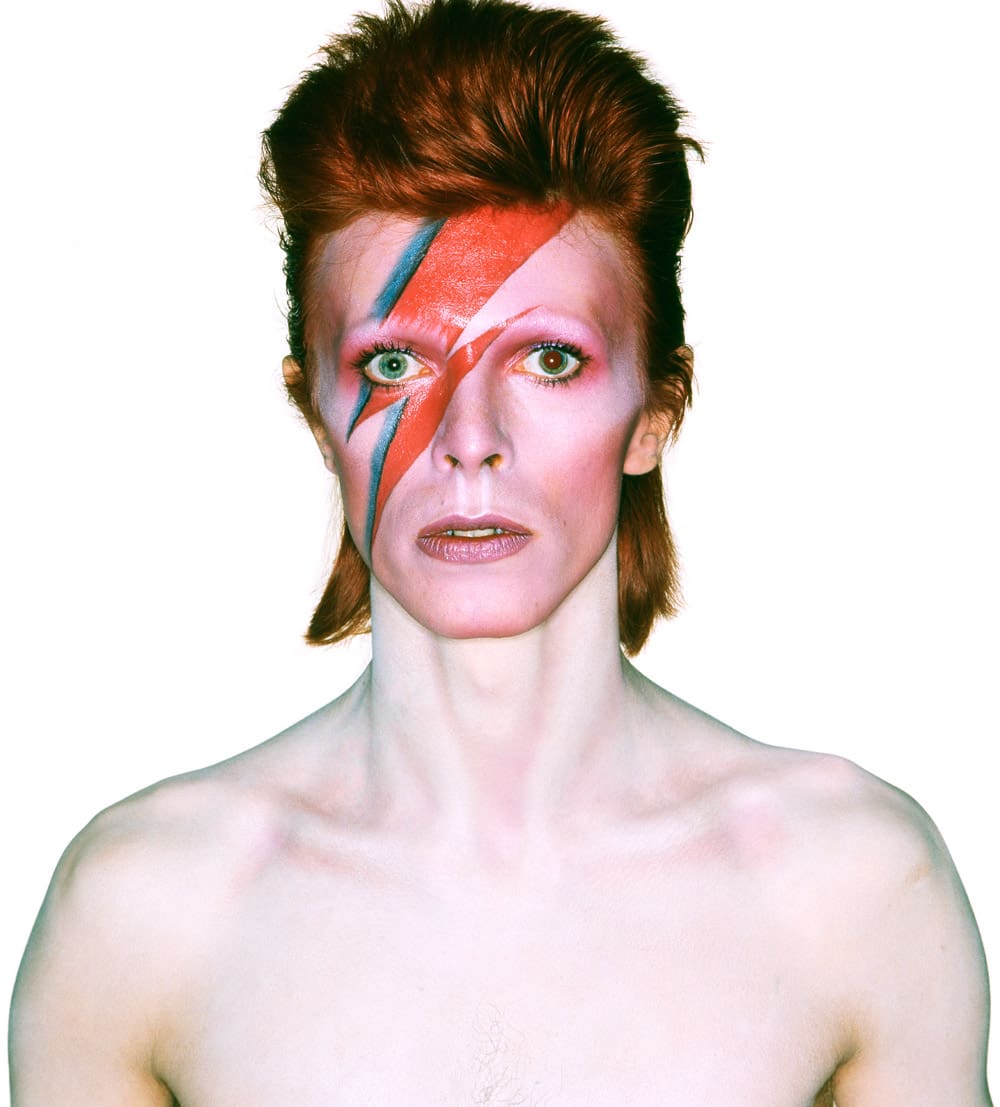A person with pale skin and bright red hair styled in a mullet poses against a white background. They have a vivid red and blue lightning bolt painted across their face and are bare-chested, looking directly at the camera.