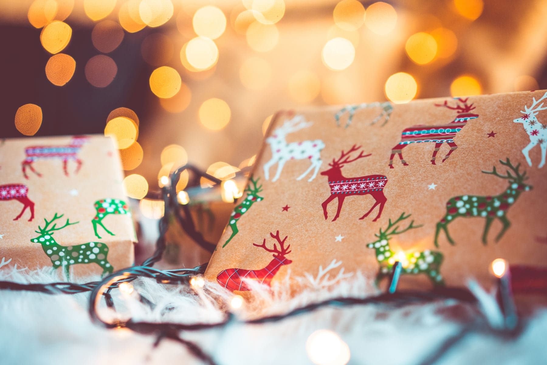 A close-up of festive gifts wrapped in paper decorated with colorful reindeer, surrounded by twinkling string lights and soft, warm bokeh lights in the background.