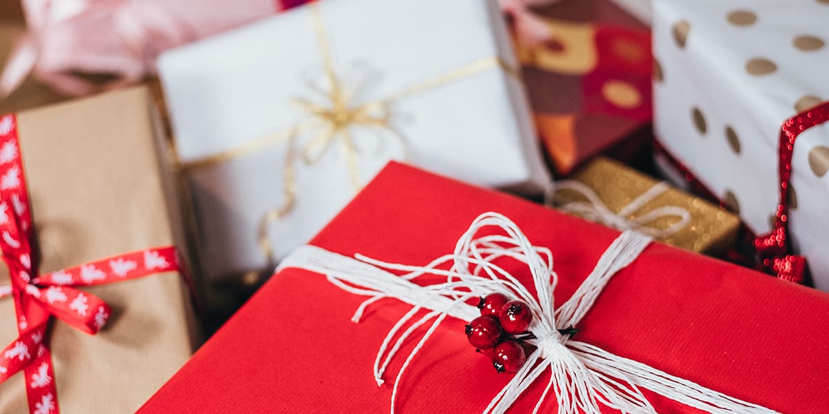 Close-up of several wrapped gifts in various colors and patterns, including red, white, gold, and brown, with different styles of ribbons and bows. One red box features a white string and festive decoration.