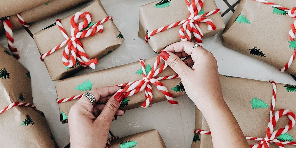 Hands with red nail polish tying a red-and-white twine bow on a brown gift wrapped with green tree prints, surrounded by several similar presents on a white surface.