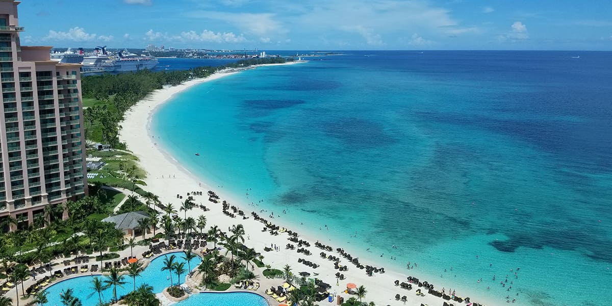 Aerial view of a tropical beach with turquoise water, white sand, palm trees, beach chairs, a swimming pool, and cruise ships in the distance under a blue sky with scattered clouds.