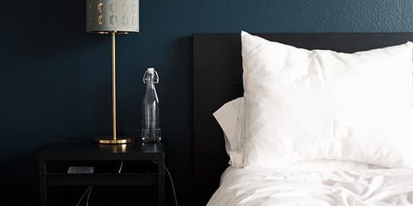 A neatly made bed with white pillows and sheets beside a black nightstand holding a lamp with a gold stand and a patterned shade, and a clear spray bottle, against a dark blue wall.
