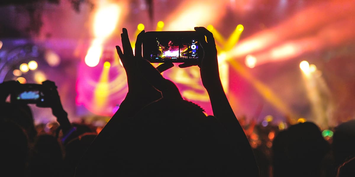 Crowd at a concert with colorful stage lights in the background; a person in the foreground is holding up a phone, recording or taking a photo of the performance.